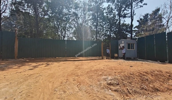 Another view of the Godrej Castillo temporary access point displaying the wide dirt path, security cabin, and tall green perimeter fencing.